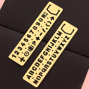 May include: Two gold-colored metal stencils with black lettering. One stencil has the numbers 1-9 and symbols. The other stencil has the alphabet.