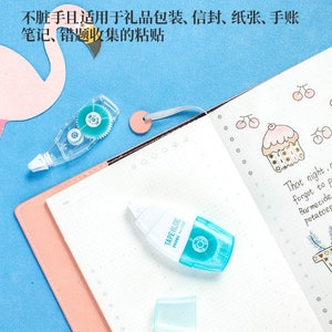 May include: Two clear tape dispensers with blue accents. The dispensers are on a pink and white notebook. The notebook has a small drawing of a cupcake with cherries on it. The text "TAPE GLUE" is visible on one of the dispensers.