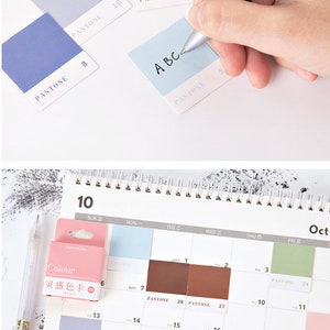 May include: A set of color swatches with the word "PANTONE" printed on each one. The swatches are in various shades of blue, gray, and brown. The swatches are arranged in a grid pattern on a white surface. A hand is writing the letters "A B C" on a light blue swatch.