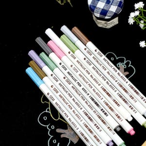 May include: A set of ten metallic pens with white barrels and black labels. The labels have the text "STA METALLIC PEN" and a number. The pens are arranged in a row from left to right, with the numbers increasing from 6551-3 to 6551-4.