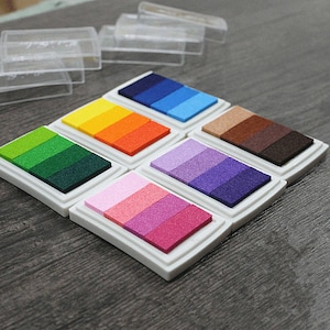 May include: Six colorful ink pads with different shades of green, orange, blue, purple, pink, and brown. Each pad has a white plastic casing and is arranged in a row on a wooden surface.