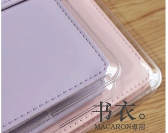 May include: Two pastel-coloured leather-bound notebooks with clear plastic covers. The top notebook is lavender and the bottom notebook is pink. The notebooks have the text "书衣。MACARON专用" printed on the front.