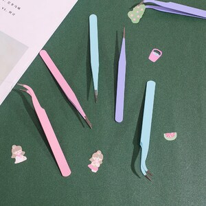 Op de afbeelding: Een set van zes pincetten in verschillende kleuren, waaronder roze, blauw en paars. De pincetten zijn gerangschikt op een groene ondergrond.