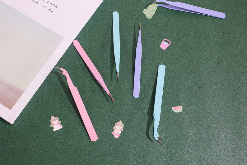 Op de afbeelding: Een set van zes pincetten in verschillende kleuren, waaronder roze, blauw en paars. De pincetten zijn gerangschikt op een groene ondergrond.