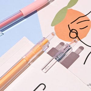 Puede incluir: Tres bol&iacute;grafos de pl&aacute;stico transparente con recargas de tinta de diferentes colores. Los bol&iacute;grafos est&aacute;n sobre una superficie blanca con un dibujo de una cara y una fruta naranja.