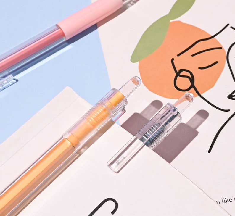 Puede incluir: Tres bol&iacute;grafos de pl&aacute;stico transparente con recargas de tinta de diferentes colores. Los bol&iacute;grafos est&aacute;n sobre una superficie blanca con un dibujo de una cara y una fruta naranja.