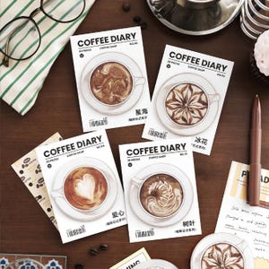 May include: Several "Coffee Diary" notepads with coffee cup illustrations and latte art designs. The notepads are white and feature text. A pen, glasses, and a striped cloth are also visible on the wooden surface.