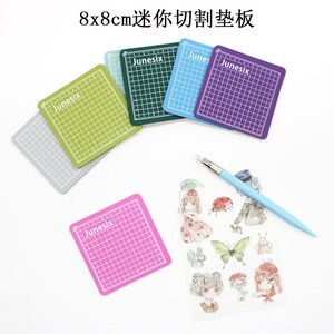May include: Six 8x8 cm self-healing cutting mats in different colors: gray, green, blue, purple, pink, and yellow. Each mat has a grid pattern and the word "Junesix" printed on it. A blue craft knife and a sheet of stickers with illustrations of a ladybug, butterfly, and girls are also shown.