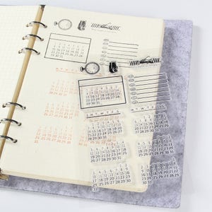Pu&ograve; includere: Timbri per calendario in acrilico trasparente con vari layout di calendario e formati di data. Include timbri con i giorni della settimana, calendari mensili e numeri di data. I timbri sono su una pagina di taccuino.