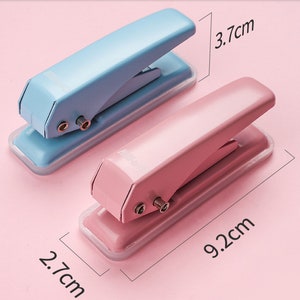 May include: Two small hole punchers, one light blue and one pink, with a clear plastic base. The punchers are 9.2 cm long and 3.7 cm wide.