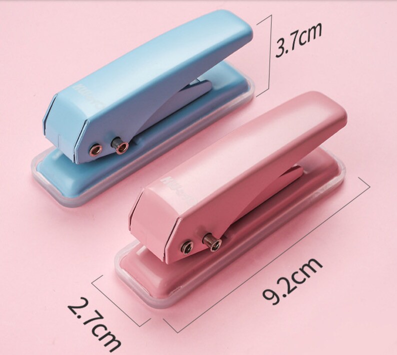 May include: Two small hole punchers, one light blue and one pink, with a clear plastic base. The punchers are 9.2 cm long and 3.7 cm wide.