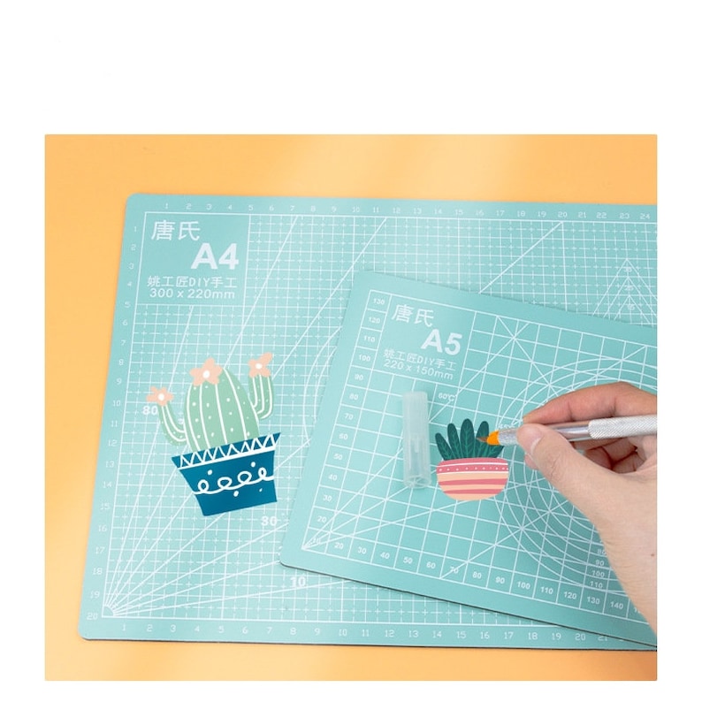 May include: Two green self-healing cutting mats with grid lines. The larger mat is labeled "唐氏 A4 姚工匠DIY手工 300 x 220mm". The smaller mat is labeled "唐氏 A5 姚工匠DIY手工 220 x 150mm". A hand is holding a craft knife and cutting a cactus sticker on the smaller mat.