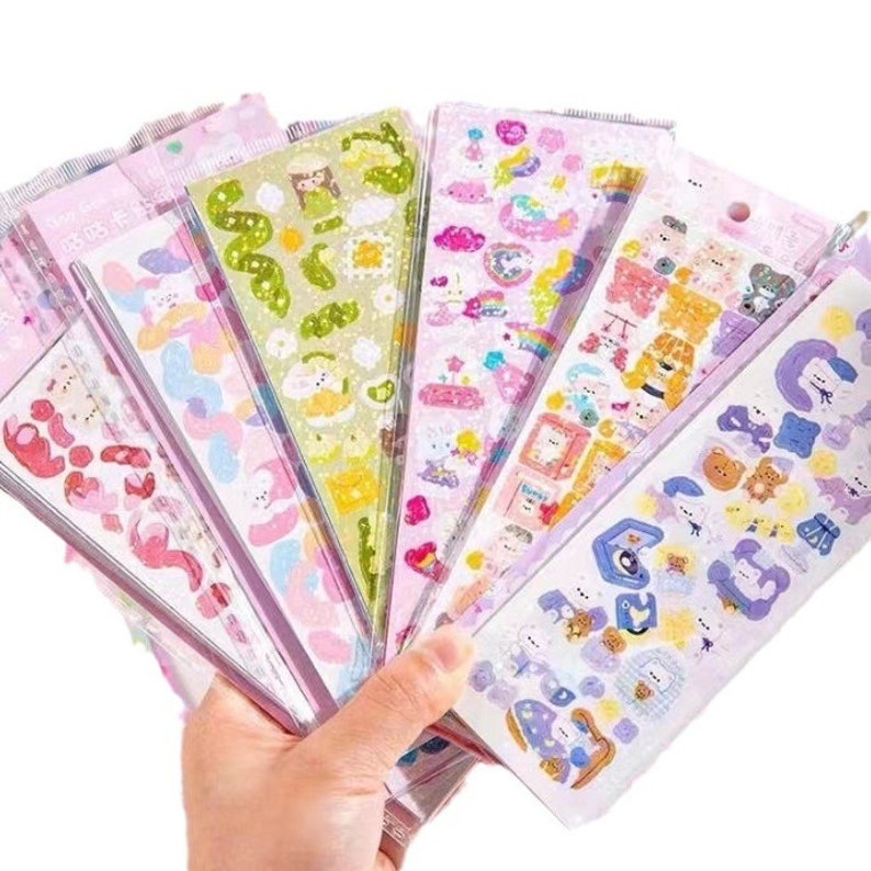 May include: A set of eight sheets of colourful stickers featuring cute cartoon characters, animals, and objects. The stickers are on a pink background with glitter accents. The stickers are perfect for decorating journals, planners, laptops, and other items.