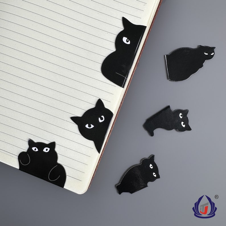 K&ouml;nnte beinhalten: F&uuml;nf schwarze Katzen-f&ouml;rmige Lesezeichen mit wei&szlig;en Augen. Die Katzen sind in verschiedenen Posen, einige schauen zur Seite, einige schauen nach vorne und einige sind zusammengerollt. Die Lesezeichen befinden sich auf einem karierten Notizbuch.