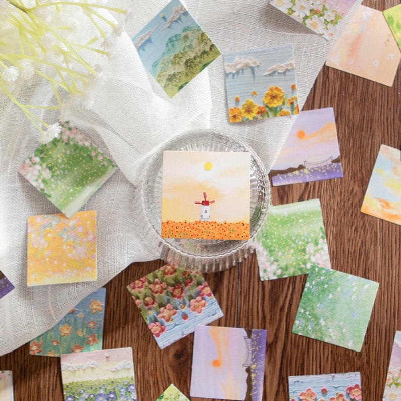 May include: Assortment of colorful square stickers with various nature scenes, including flowers, a windmill, a bridge, and a sunset. The stickers are arranged on a wooden surface with a white cloth in the background.