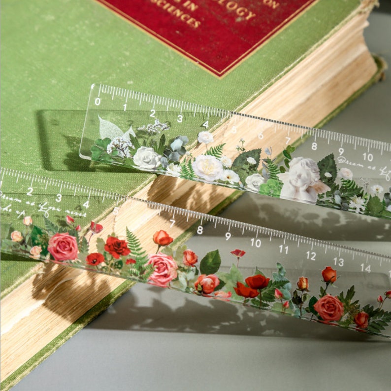 May include: Two clear plastic rulers with floral designs. The top ruler has white flowers and green leaves. The bottom ruler has red flowers and green leaves. Both rulers have black numbers printed on them.