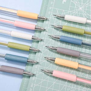 Puede incluir: Un conjunto de diez l&aacute;pices mec&aacute;nicos con cuerpos transparentes y agarres de colores. Los colores son amarillo, rosa, blanco, verde, azul, gris y naranja. Los l&aacute;pices est&aacute;n dispuestos en una fila sobre una estera de corte verde con una regla marcada en cent&iacute;metros.