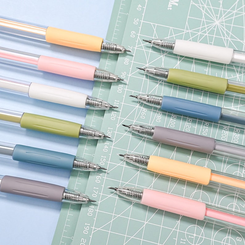 Puede incluir: Un conjunto de diez l&aacute;pices mec&aacute;nicos con cuerpos transparentes y agarres de colores. Los colores son amarillo, rosa, blanco, verde, azul, gris y naranja. Los l&aacute;pices est&aacute;n dispuestos en una fila sobre una estera de corte verde con una regla marcada en cent&iacute;metros.