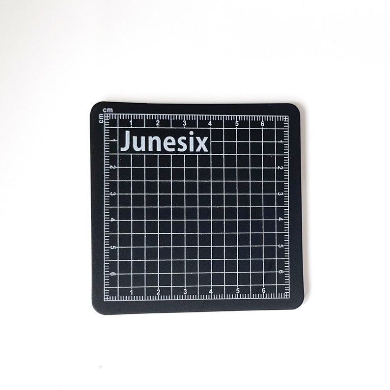 May include: Black self-healing cutting mat with a grid pattern and centimeter markings. The mat has the word "Junesis" printed in white on the top left corner.