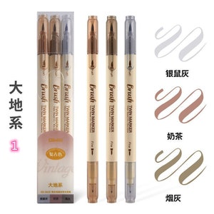 May include: A set of vintage-style twin marker brush pens in shades of brown, gold, and silver. The pens are labeled "Brush Twin Marker" and "Fine". The packaging includes Chinese text and color swatches.
