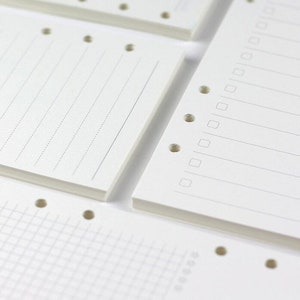 Op de afbeelding: Close-up van een set witte papieren notitieboek-navullingen met geperforeerde gaten. De navullingen hebben verschillende papierstijlen, waaronder gestreept, geruit en blanco.