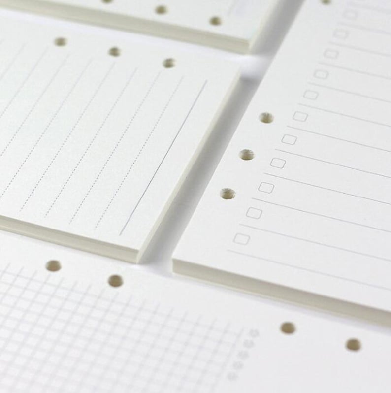Op de afbeelding: Close-up van een set witte papieren notitieboek-navullingen met geperforeerde gaten. De navullingen hebben verschillende papierstijlen, waaronder gestreept, geruit en blanco.