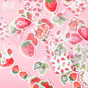 K&ouml;nnte beinhalten: Eine Blatt mit bunten Aufklebern mit Erdbeeren in verschiedenen Designs. Die Aufkleber befinden sich auf einem rosa Hintergrund mit einem wei&szlig;en Netz&uuml;berzug. Die Aufkleber enthalten Erdbeeren in K&ouml;rben, Erdbeeren mit Bl&auml;ttern und gemusterte Erdbeeren.