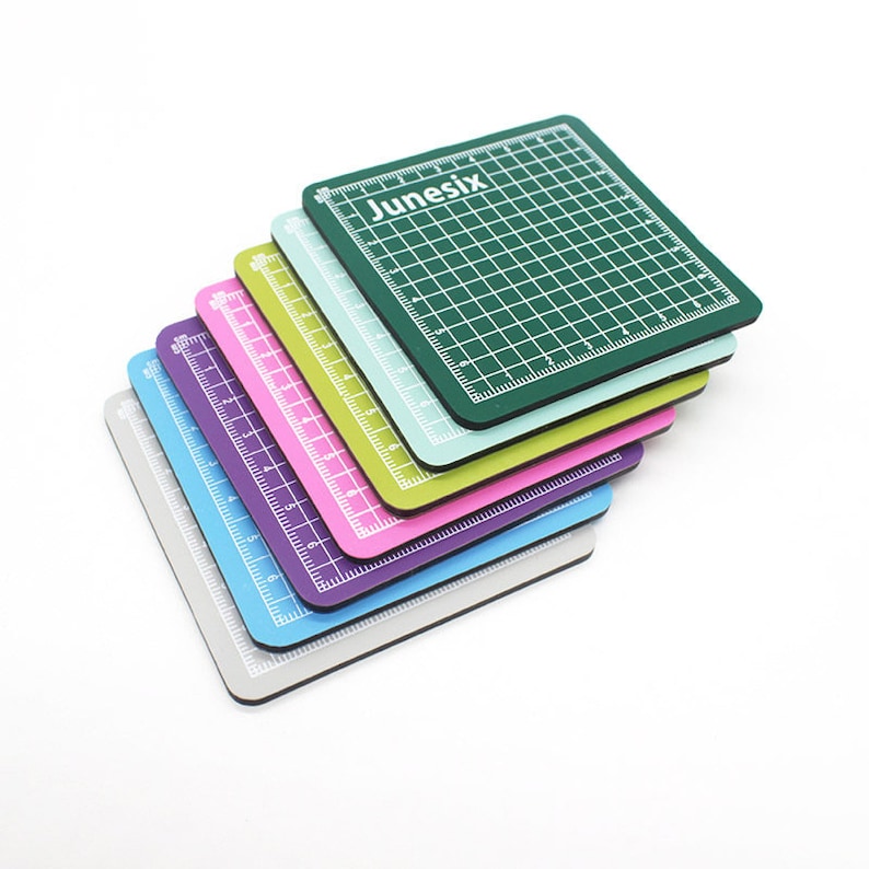 May include: A stack of six self-healing cutting mats in various colors, including green, light blue, purple, pink, and gray. The top mat has the word "Junesix" printed on it.