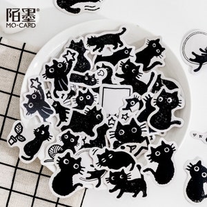 May include: A sheet of black and white stickers featuring various illustrations of cats. The stickers are on a white plate with a black and white checkered background. The text "MO-CARD" is visible in the top left corner of the image.