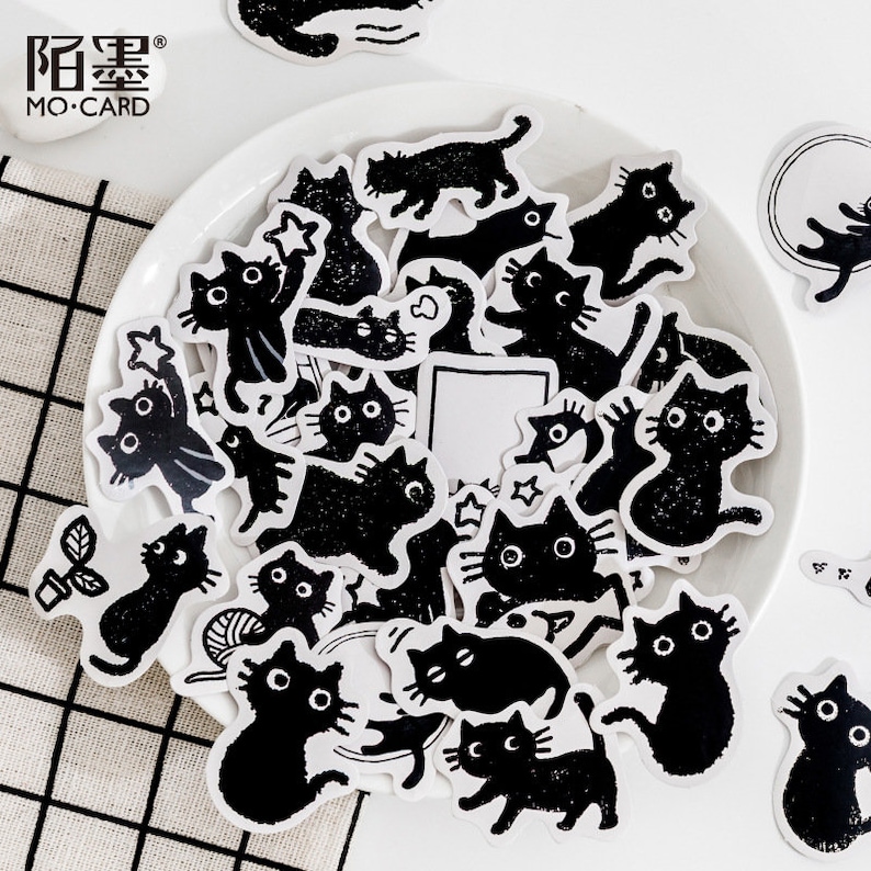 May include: A sheet of black and white stickers featuring various illustrations of cats. The stickers are on a white plate with a black and white checkered background. The text "MO-CARD" is visible in the top left corner of the image.