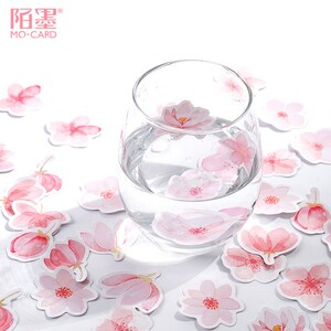 May include: Pink watercolor flower stickers scattered on a white surface with a glass of water in the center. The stickers feature delicate pink flowers with white centers.