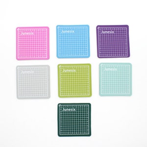 May include: Set of eight self-healing cutting mats in various colors, including pink, blue, purple, gray, green, light blue, dark green, and black. Each mat features a grid pattern and the word "Junesix" printed in white.