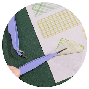 Op de afbeelding: Twee pincetten, een blauwe en een zilveren, worden gebruikt om een ​​vel gepatenteerd papier te verwijderen. Het papier heeft een geruit patroon in groen, geel en wit.