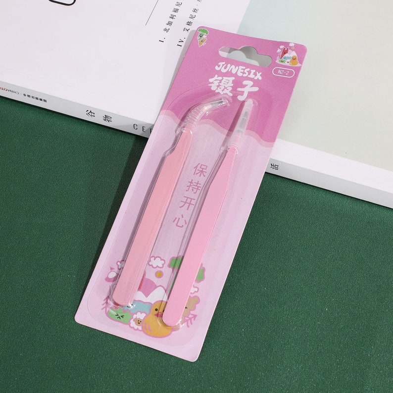 Op de afbeelding: Een roze plastic verpakking met twee roze pincetten. De verpakking heeft een cartoonontwerp met een gele eend en andere dieren. De tekst op de verpakking luidt "JUNESIX 镊子 保持开心 2-ZN".