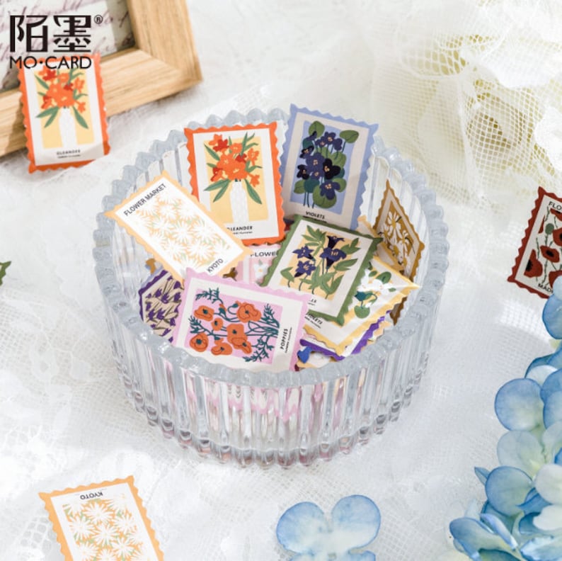 May include: A glass bowl filled with colorful floral patterned stickers. The stickers are shaped like postage stamps and feature different types of flowers, including violets, poppies, and daisies. The text on the stickers includes the names of the flowers and the words "Flower Market".