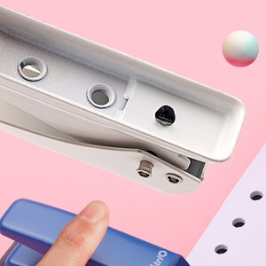 May include: A white metal punch with a black handle and a hole punched through a piece of paper. The punch is on a pink surface.