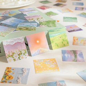 May include: A set of colorful square stickers featuring various nature scenes, including flowers, mountains, and skies. The stickers are arranged on a white surface and a green box.