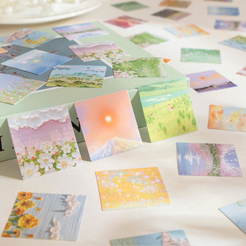 May include: A set of colorful square stickers featuring various nature scenes, including flowers, mountains, and skies. The stickers are arranged on a white surface and a green box.