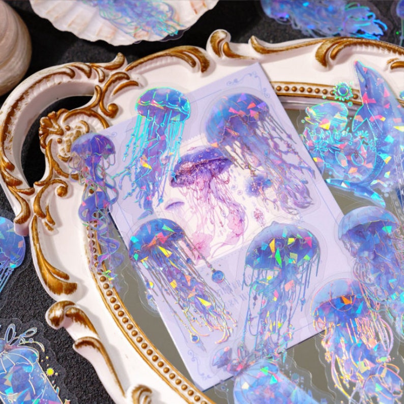 May include: A sheet of iridescent holographic stickers featuring jellyfish in various poses. The stickers are on a white background with a light blue and purple color scheme.