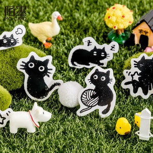 May include: A set of black and white cat stickers with different poses, including a cat sleeping, a cat playing with yarn, and a cat sitting. The stickers are on a green grass background with a small white duck and a yellow chick.