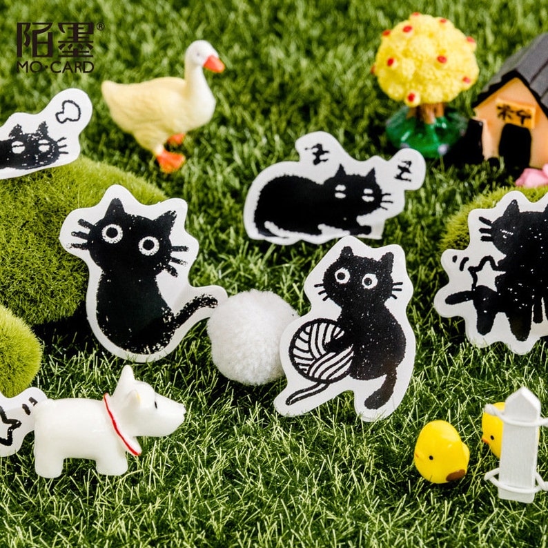 May include: A set of black and white cat stickers with different poses, including a cat sleeping, a cat playing with yarn, and a cat sitting. The stickers are on a green grass background with a small white duck and a yellow chick.