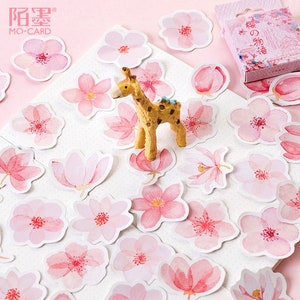 May include: A sheet of pink and white watercolor stickers featuring various floral designs, including cherry blossoms and magnolia flowers. A small giraffe figurine is placed on the sheet.