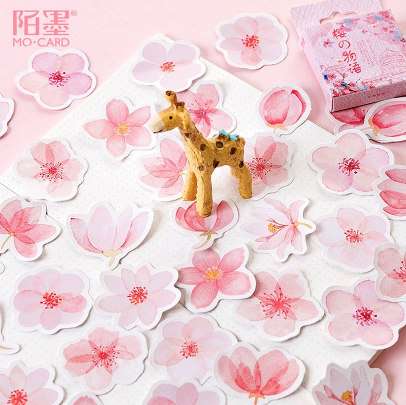 May include: A sheet of pink and white watercolor stickers featuring various floral designs, including cherry blossoms and magnolia flowers. A small giraffe figurine is placed on the sheet.