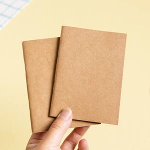 Könnte beinhalten: Zwei kleine, leere Notizbücher mit braunen Umschlägen werden in einer Hand gehalten. Die Notizbücher sind rechteckig und haben ein schlichtes, minimalistisches Design. Der Hintergrund ist hellgelb.