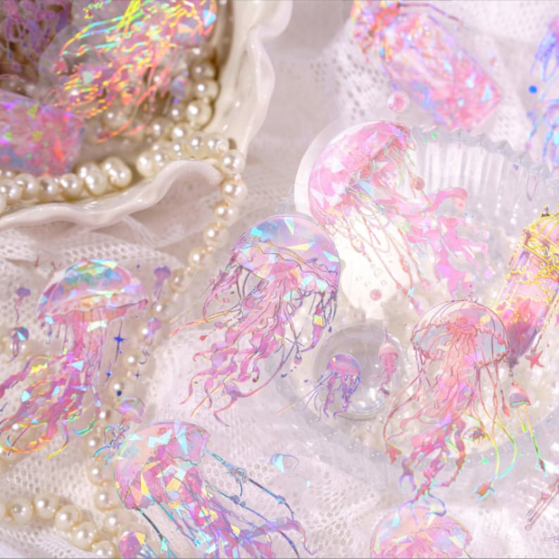 May include: A collection of iridescent holographic jellyfish stickers in various sizes and poses. The stickers are pink, purple, and blue with a glittery finish. The stickers are on a white background with a pearl necklace.