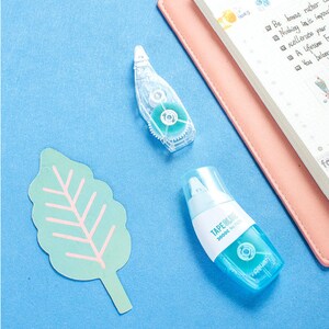 May include: Two blue and white correction tape dispensers on a light blue surface. The dispensers are labelled "TAPE BLADE" and "№7305".