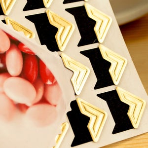 May include: Gold and black corner stickers in a chevron pattern. The stickers are designed to be used in a scrapbook or photo album.