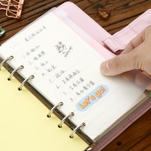May include: A yellow and pink planner with a clear plastic cover. The planner has a ring binding and is open to a page with a to-do list written in Chinese. The list includes items like "practice yoga" and "go swimming". There is a sticker on the page that says "Let's go!"