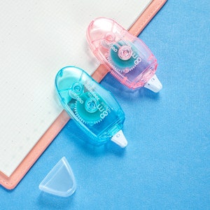 May include: Two transparent tape dispensers, one blue and one pink, with a white tape dispenser lid on a blue surface. The dispensers have a clear plastic body and a white tape dispenser lid. The dispensers have the text "Tombow Tape" and "Zero" printed on them.
