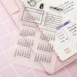 Pu&ograve; includere: Timbri calendario in acrilico trasparente con vari layout mensili e settimanali. I timbri includono numeri, giorni della settimana ed elementi decorativi. I timbri sono su uno sfondo rosa.